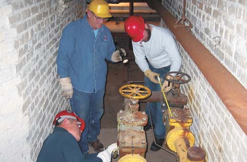 Glenn Bear, center, and Charlie Griesen, right, of Utility Services help measure the valve of a steam pipe that runs...